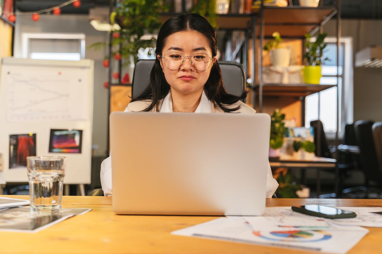 Asian woman with eyeglasses working on a laptop in a modern office setting, surrounded by charts and plants.